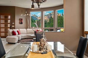 Ein modernes Wohnzimmer mit einer Couch, einem Esstisch mit Vase und Kerzen sowie großen Fenstern mit Blick auf die Berge.