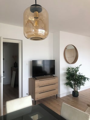 Ein modernes Wohnzimmer mit Flachbildfernseher auf einem Holzschrank, einer Pendelleuchte, einem runden Spiegel und einem Blumentopf.