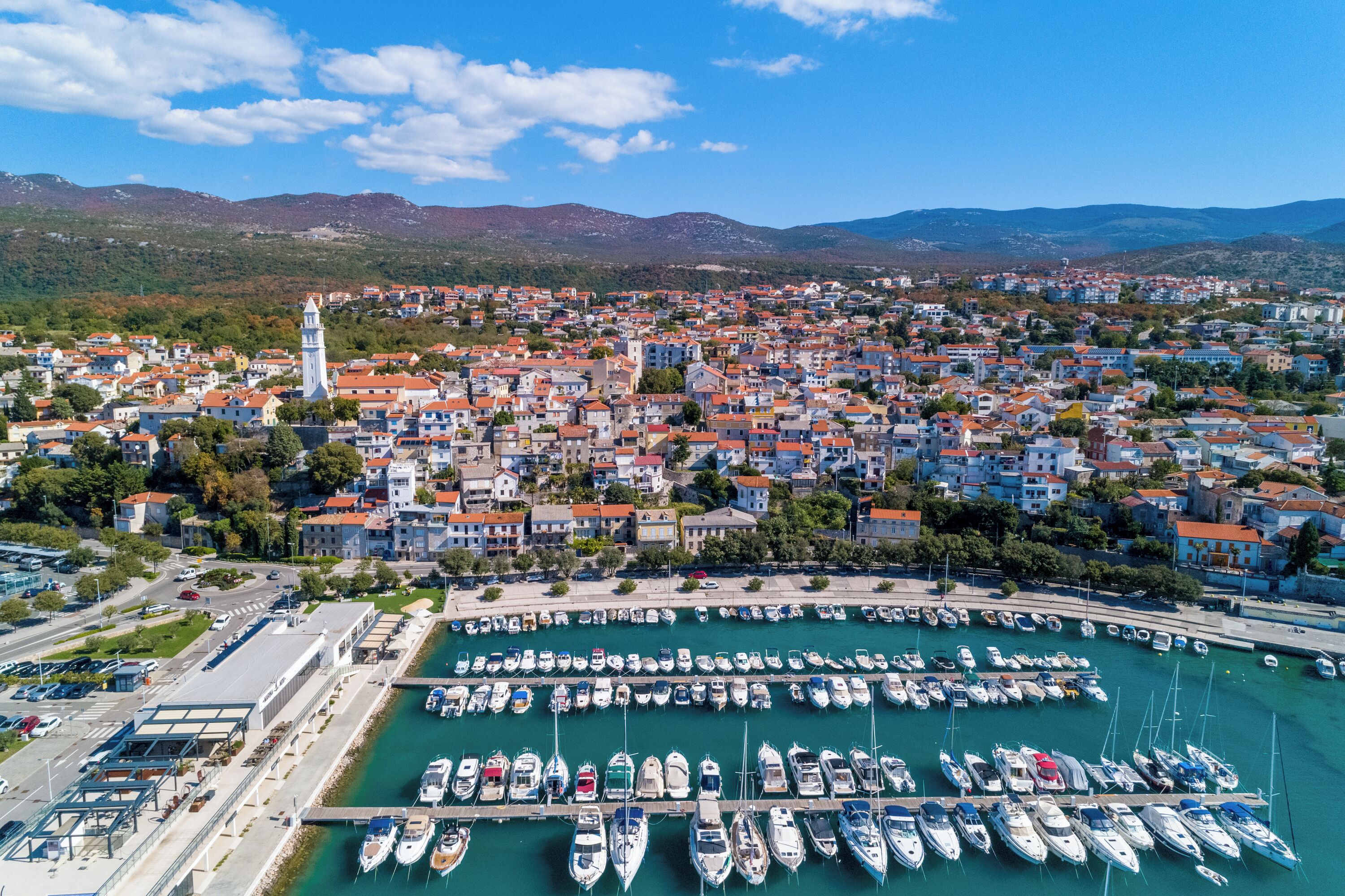 A coastal town with a marina, buildings with red roofs, and a prominent tower.