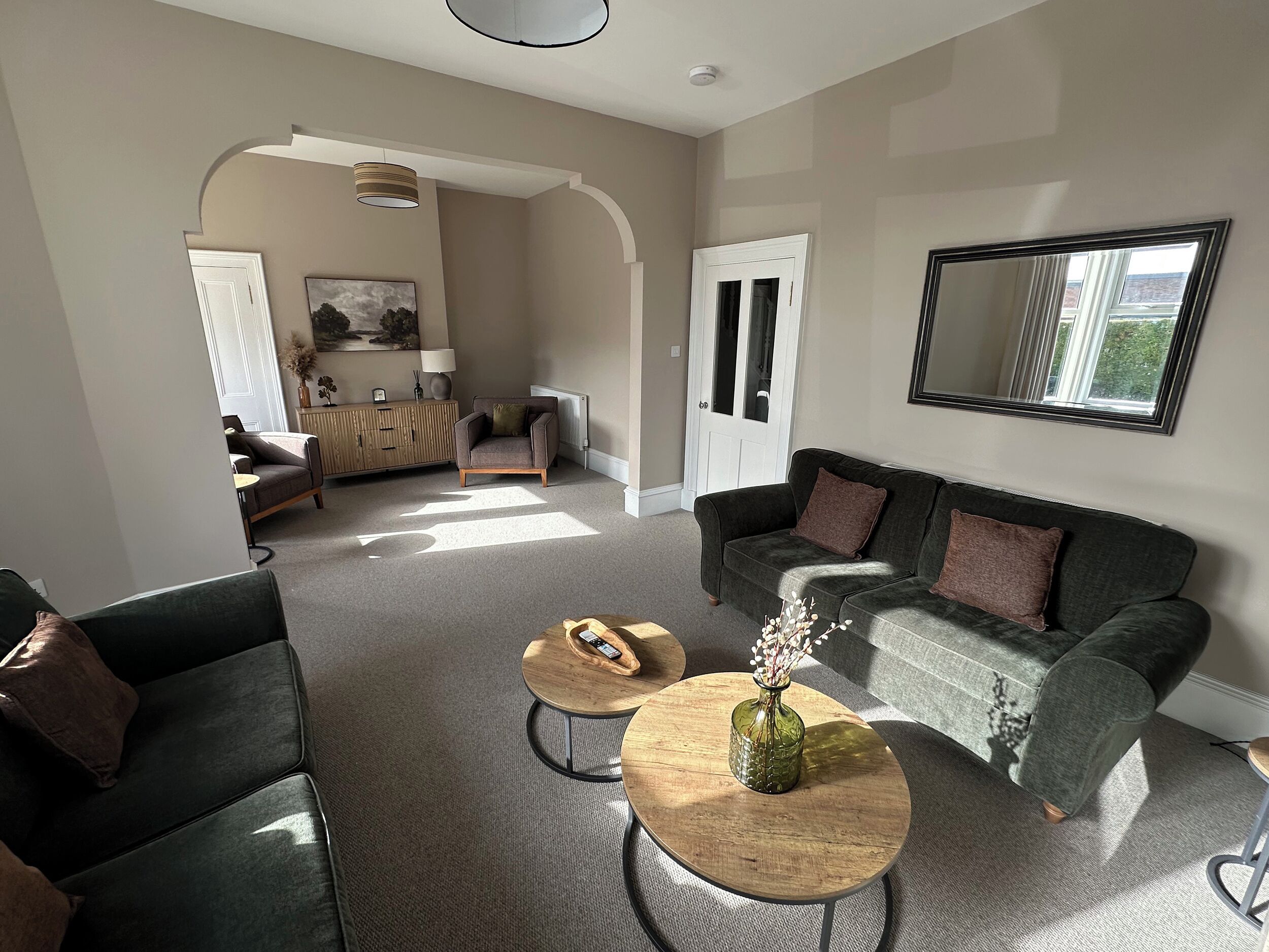 A modern living room with a grey sofa, a round wooden coffee table, and a large mirror on the wall.