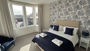 Ein Schlafzimmer mit einem großen Bett, einem Nachttisch, einem Sessel und einem Fenster mit Vorhängen.