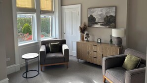 Ein Zimmer mit einer Couch, einem kleinen runden Tisch, einem Schrank, einem Gemälde und einem Fenster mit Jalousien.