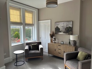 A room with a sofa, a small round table, a cabinet, a painting, and a window with blinds. - Abbotsford (Kelso)