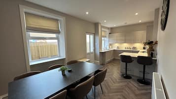 A modern kitchen with a dining area, wooden herringbone flooring, and a large window with blinds.