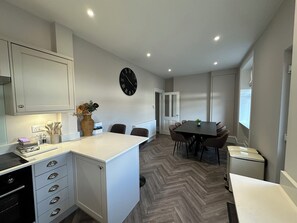 A modern kitchen with white cabinetry, a dining area with a table and chairs, a clock on the wall, and a herringbone-patterned floor. - Abbotsford (Kelso)