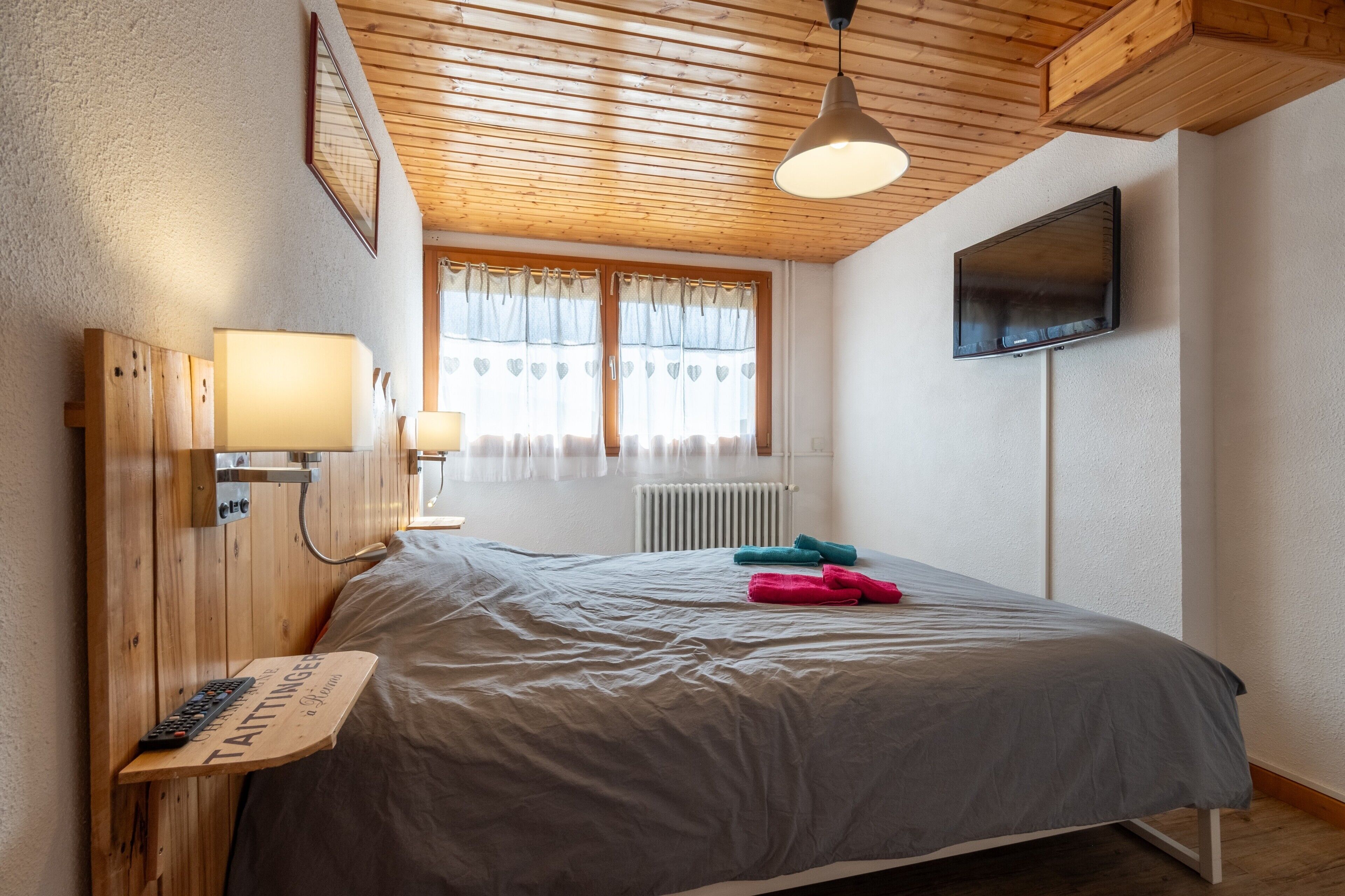 A bedroom with a wooden headboard, a bedside table with a lamp, a bed with a grey blanket, a TV on the wall, and a window with curtains.
