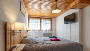 A bedroom with a wooden headboard, a bedside table with a lamp, a bed with a grey blanket, a TV on the wall, and a window with curtains. - Apartment 'Les Airelles' with Mountain View, Private Terrace and Wi-Fi (La Bresse)