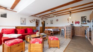 A spacious living area with a red sofa, wooden ottomans, a dining table, and a kitchen area with wooden cabinets and a red refrigerator. - Apartment 'Les Airelles' with Mountain View, Private Terrace and Wi-Fi (La Bresse)