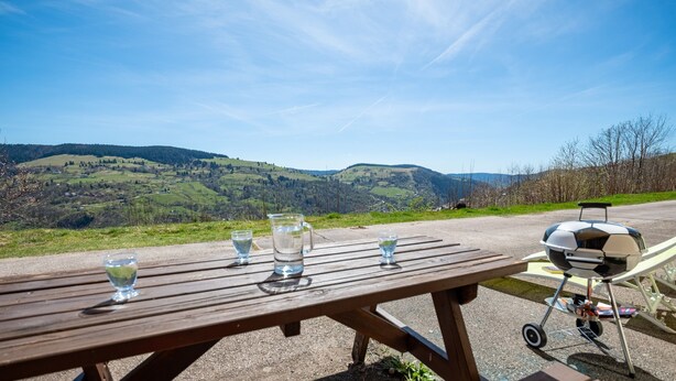 A picnic table with a grill and glasses of water, overlooking a hilly landscape. - Apartment 'Les Airelles' with Mountain View, Private Terrace and Wi-Fi (La Bresse)