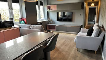 A modern kitchen with a dining area, a flat-screen TV mounted above the seating area, and a large window providing natural light.