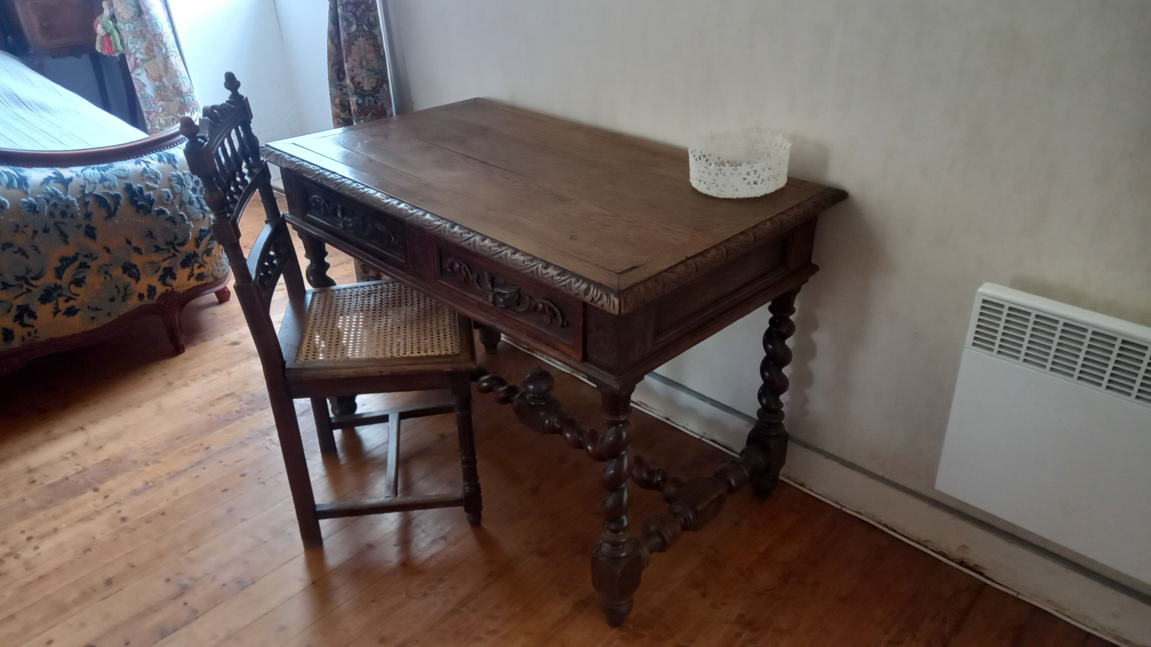 A wooden table with intricate carvings, a chair with a woven seat, and a white radiator against a plain wall.