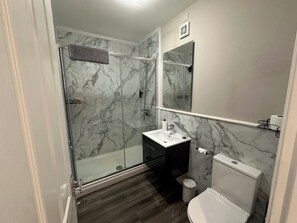 A bathroom with a glass-enclosed shower, a white sink, a toilet, and a marble-patterned wall. - The Esk (Melrose)