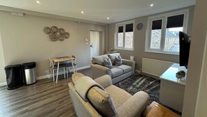 A living room with a sofa, a coffee table, a dining table with chairs, a radiator, and a window with a view of buildings. - The Esk (Melrose)