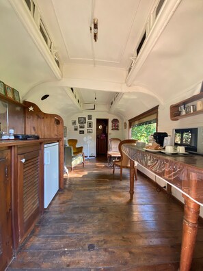 A narrow, wooden-paneled interior with a kitchen area, dining chairs, and a small refrigerator.