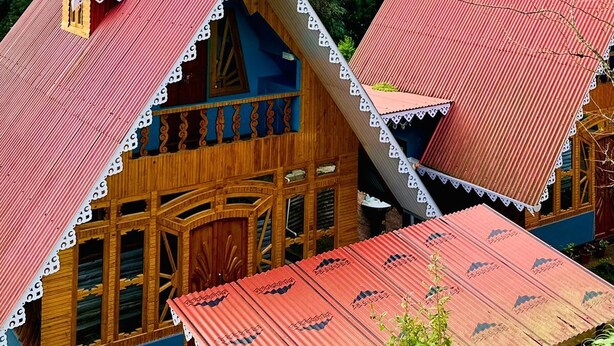 A traditional wooden house with a red roof, surrounded by lush greenery and mountains. - Ganganiwas Homestay Sonada (Darjeeling)