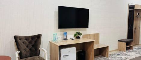 A compact room with a single bed, a bedside table, a TV mounted on the wall, and a small refrigerator.