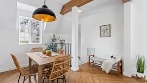 Een eetgedeelte met een houten tafel, rieten stoelen, een hangende hanglamp, een bank en een potplant.