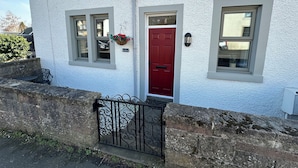 A white building with a red door, two windows, a hanging flower basket, and a black wrought iron gate. - Pergola (Melrose)