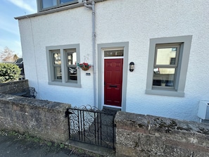 A white building with a red door, two windows, a hanging flower basket, and a black wrought iron gate. - Pergola (Melrose)