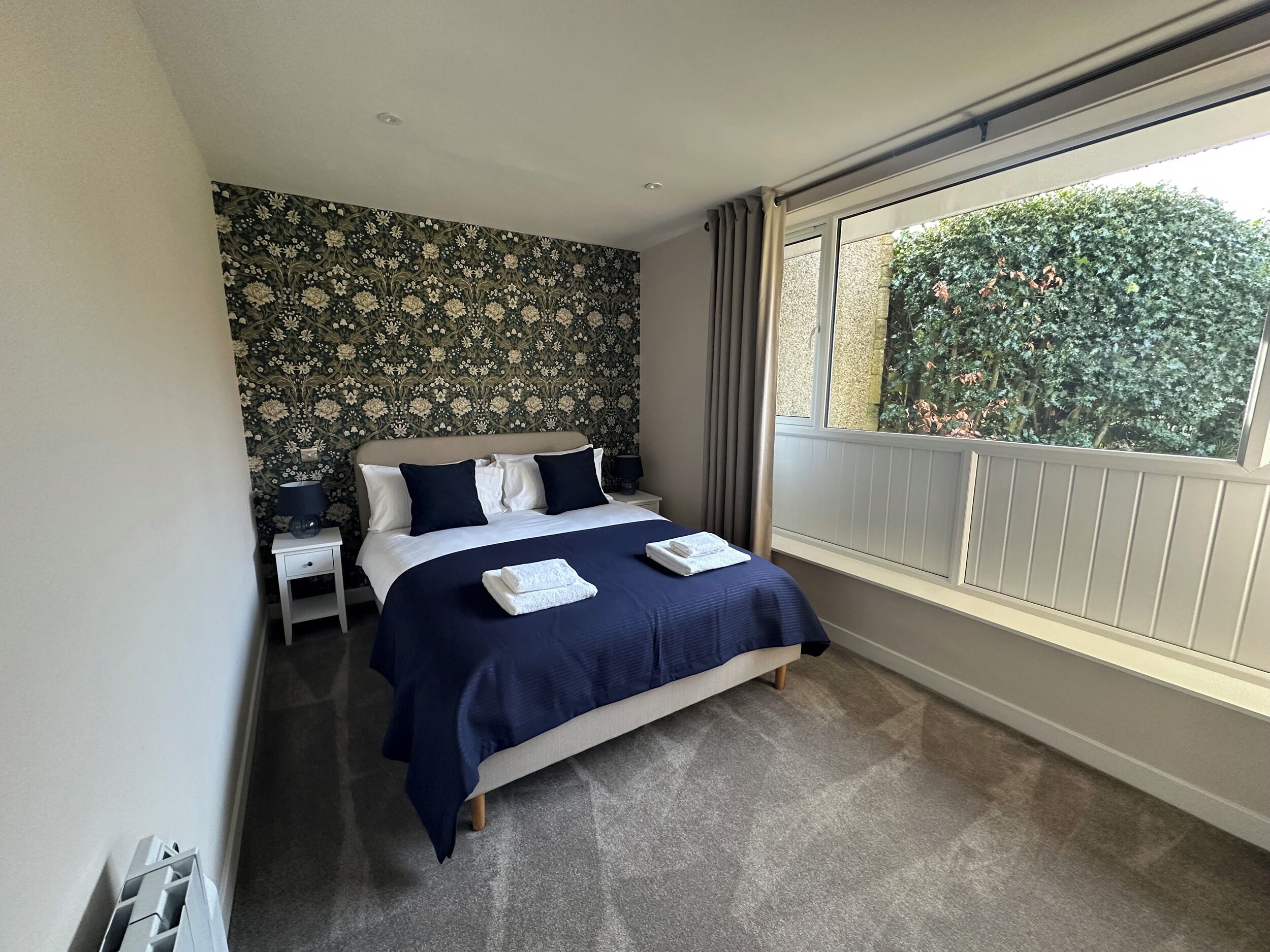 A bedroom with a bed, a nightstand, a window with a view of greenery, and a patterned wallpaper.