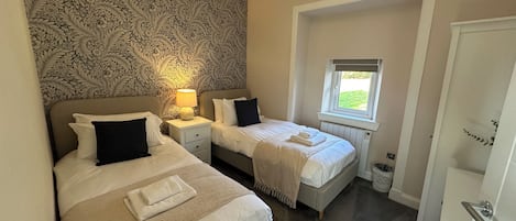 A bedroom with two beds, a patterned wallpaper, a window with a view, and a ceiling light.