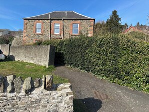 Una casa de piedra con dos ventanas, un camino de entrada pavimentado y un muro de piedra.