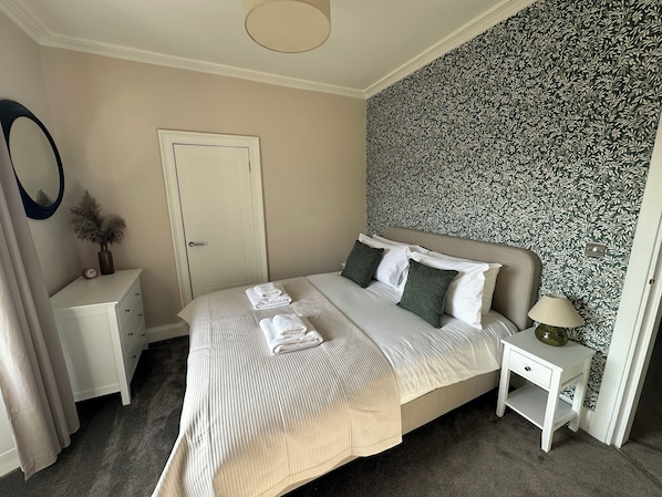 A neatly made bed with pillows and folded towels, a bedside table with a lamp, a dresser, a door, and a patterned wallpaper. - Broombank (Melrose)
