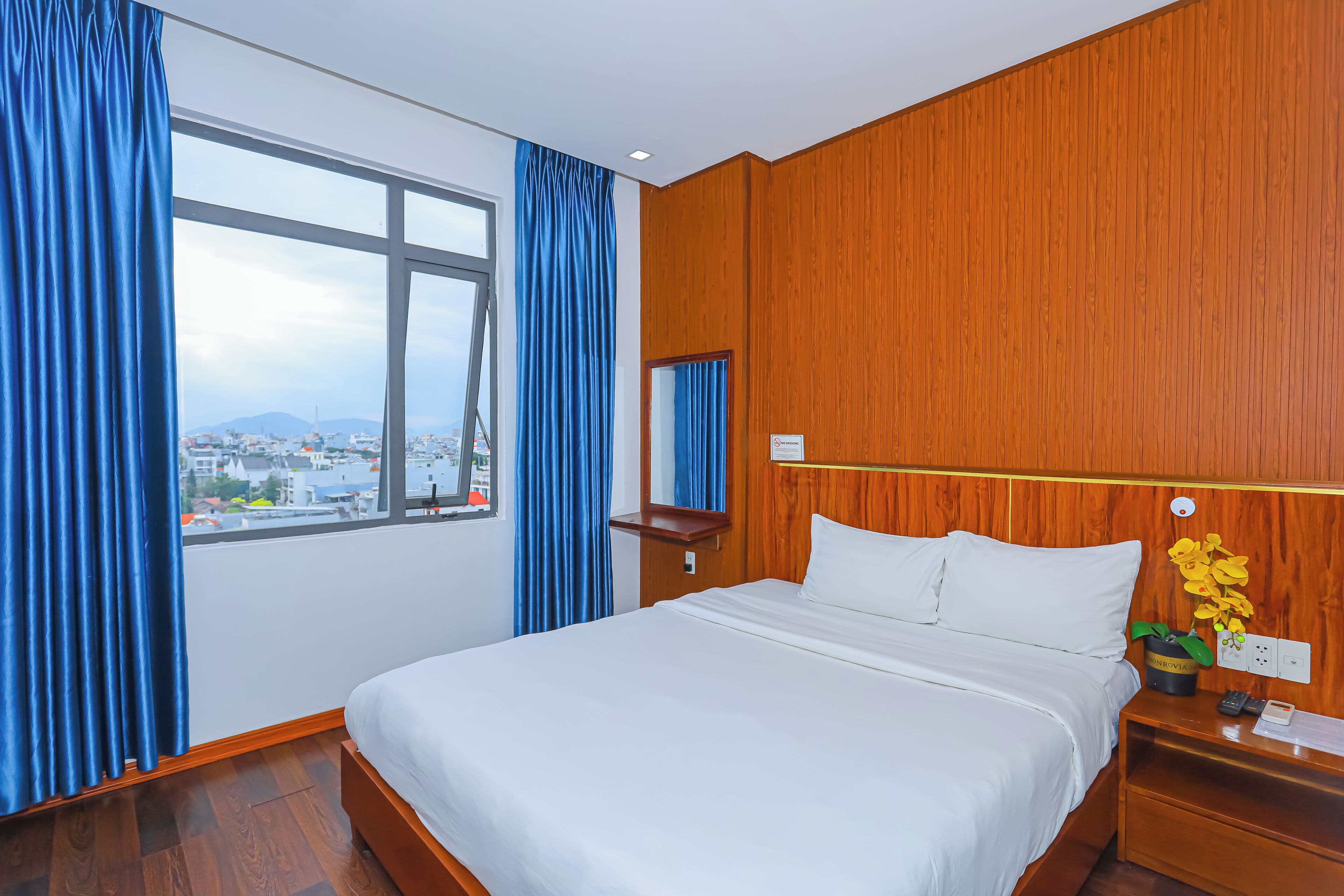 A hotel room with a large bed, a wooden headboard, a nightstand, a mirror, and a view of the city through a large window.
