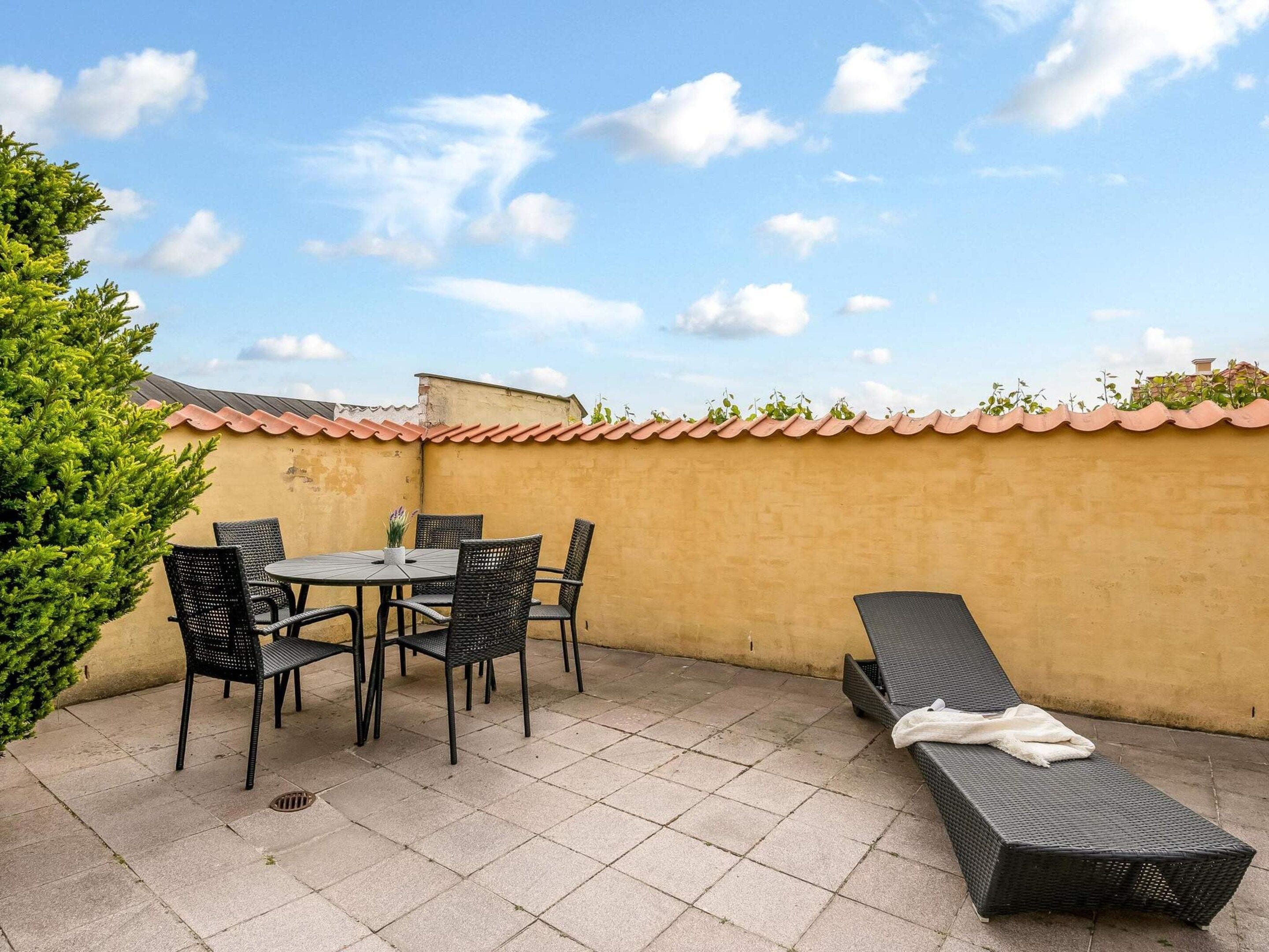 Un patio con una mesa, sillas, una tumbona y una pequeña planta en maceta.