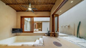 Un interior moderno con un televisor de pantalla plana, ventilador de techo y un área de estar que conduce a un espacio exterior con fuente de agua y plantas de cactus.