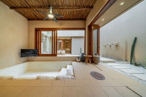 A modern interior with a flat-screen TV, a ceiling fan, and a seating area leading to an outdoor space with a water feature and cactus plants. - Uluwatu Rooftop Villa with Private Pool (South Kuta)