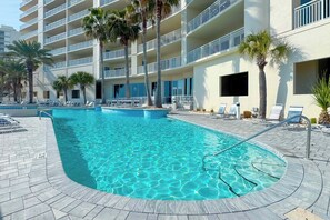 A swimming pool with lounge chairs and palm trees, surrounded by a multi-story building with balconies. - Admirals Quarters 708 | Beachfront Views | Luxury Amenities | Heart of Orange Beach (Orange Beach)