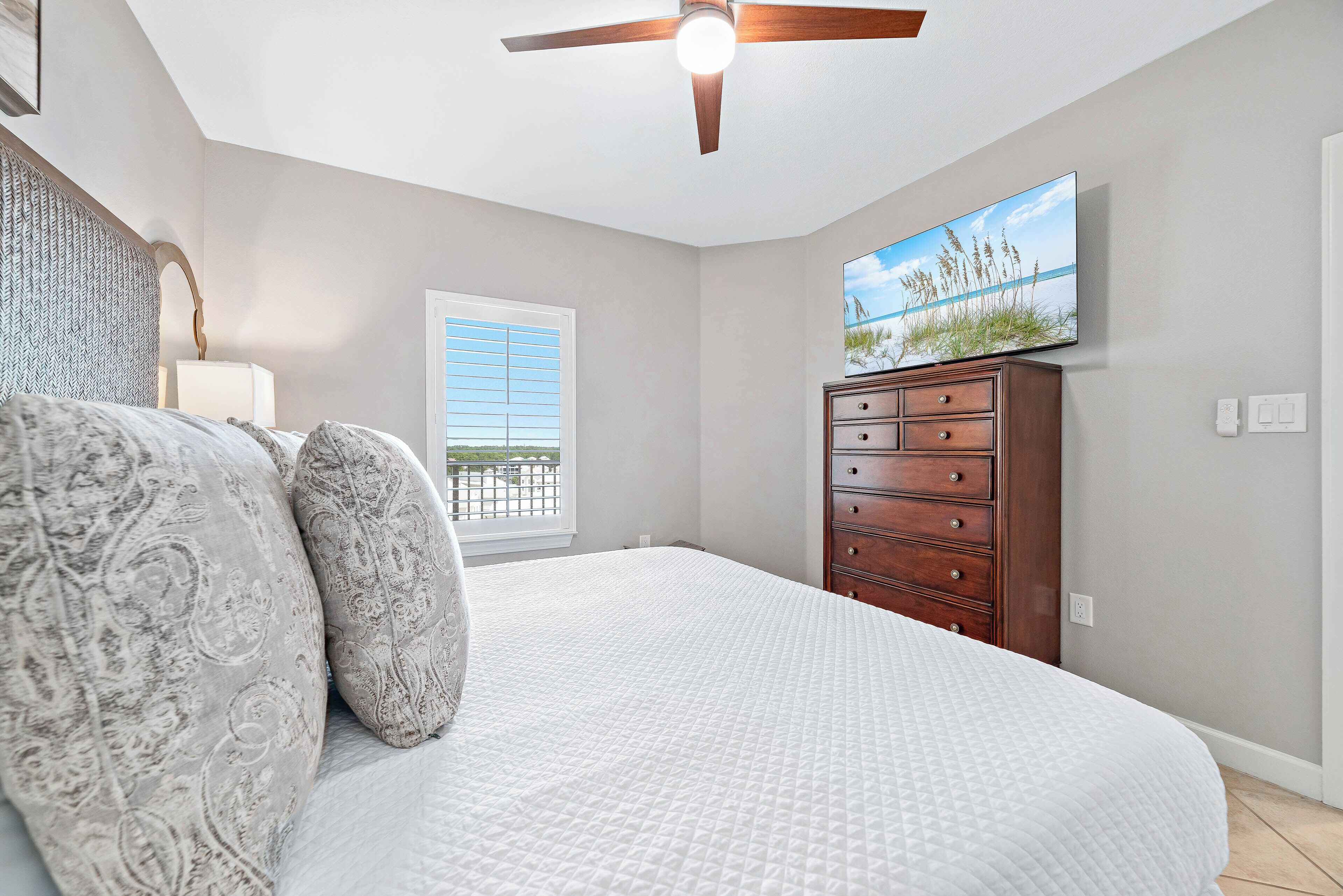 A bedroom with a bed, a dresser, a ceiling fan, a window with blinds, and a wall-mounted TV.