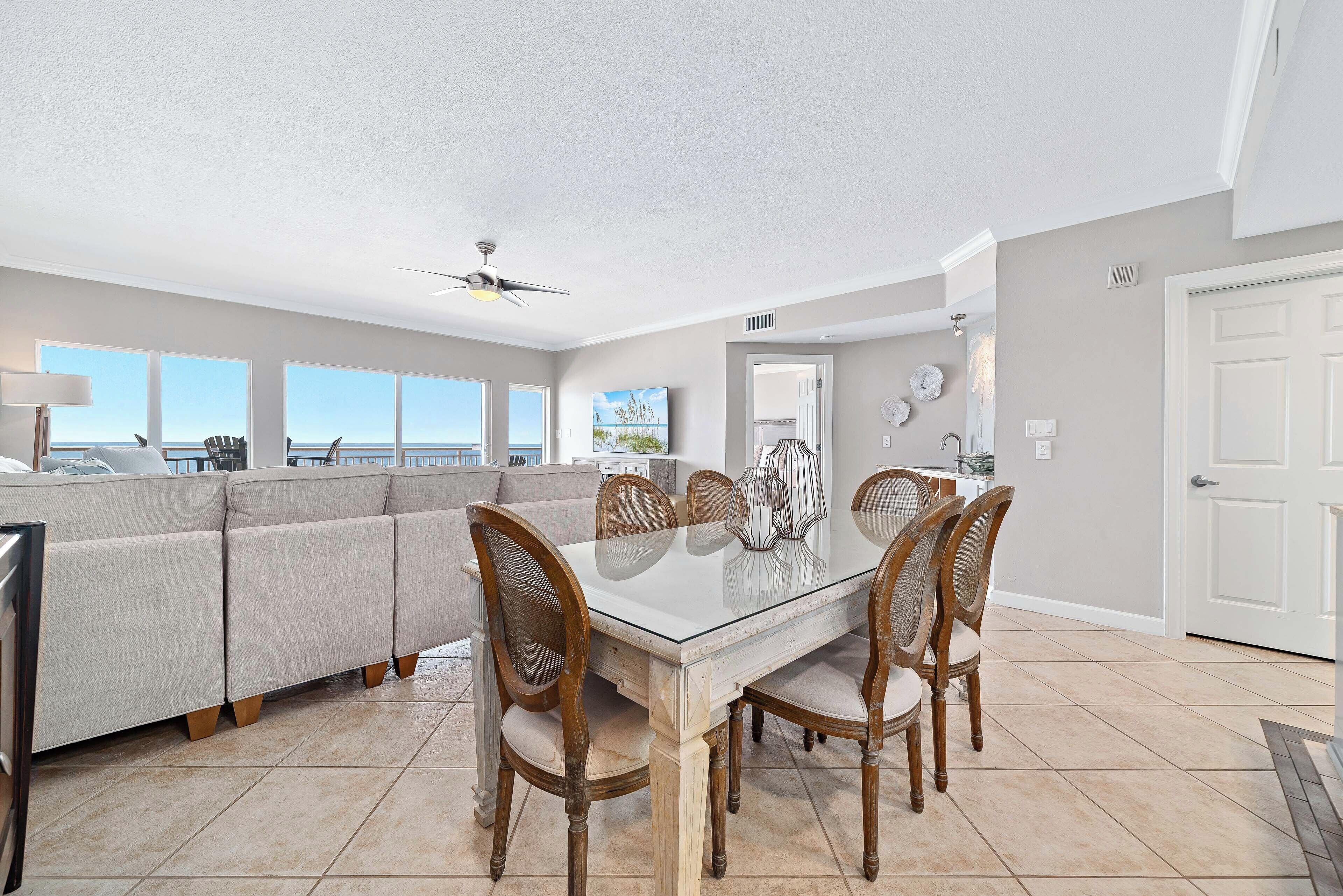 A dining area with a glass table, wooden chairs, and a view of the ocean through large windows.