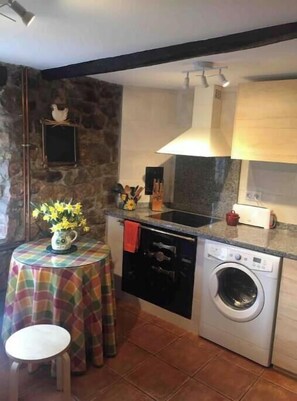 Une cuisine compacte avec un mur en pierre, un lave-linge, une cuisinière et une petite table à manger recouverte d’une nappe colorée.