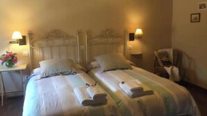 A double bed with two pillows, a bedside table with a lamp, and a chair in a room with wooden ceiling beams. - Villafañe House (Brañosera)