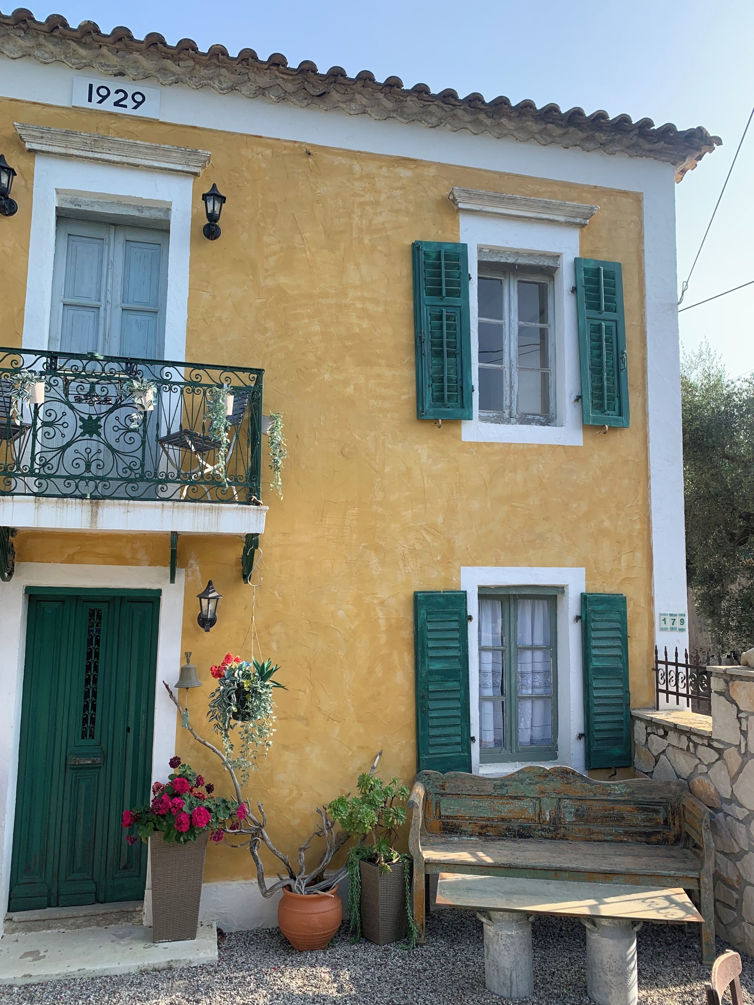 A yellow building with green shutters, a balcony with wrought iron railings, and a stone bench.
