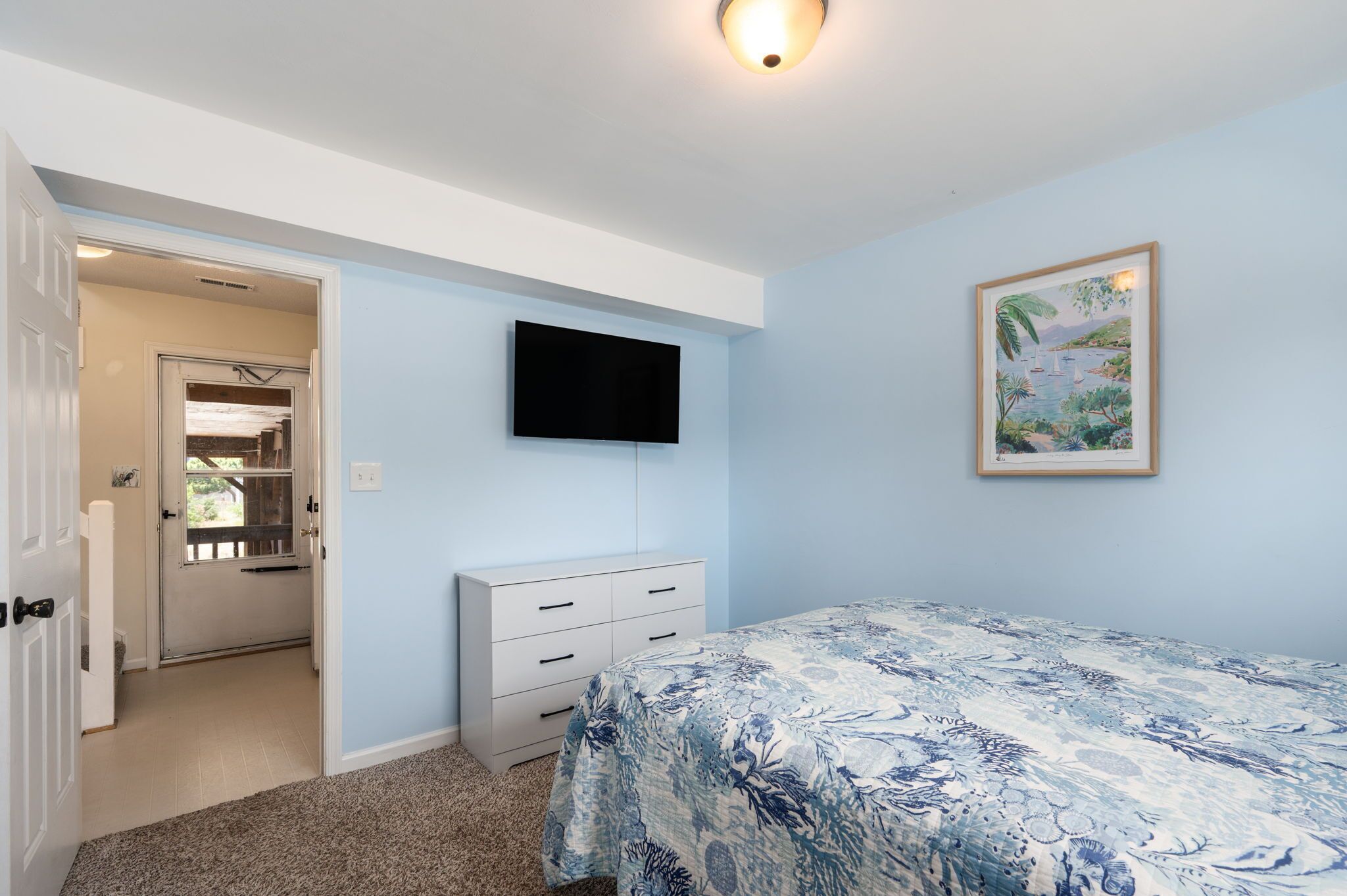 Une chambre à coucher avec un lit, un téléviseur fixé au mur, une porte donnant sur une autre pièce et un tableau encadré accroché au mur.