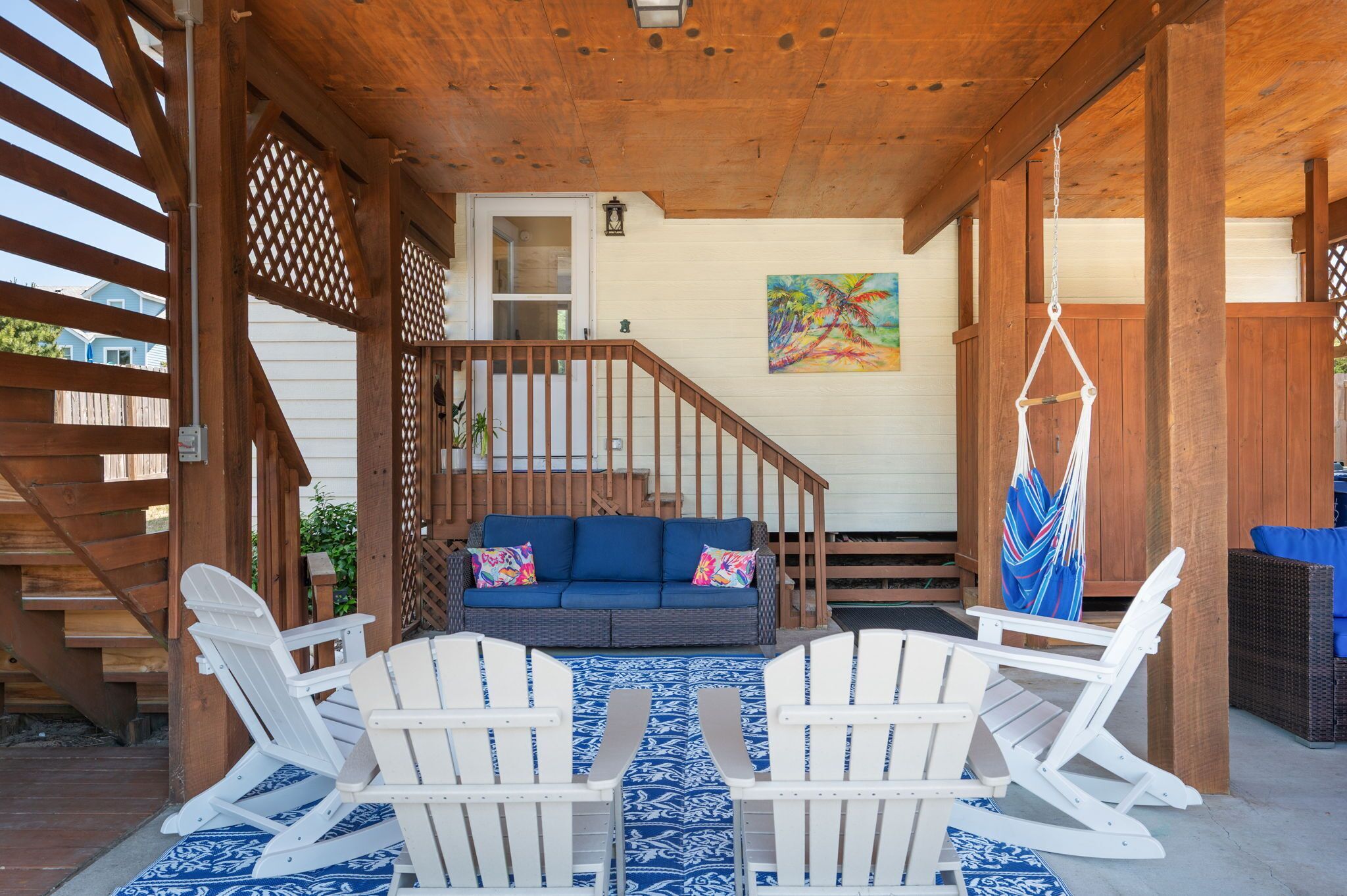A covered outdoor area with a wooden staircase, a blue sofa with colorful cushions, white Adirondack chairs, and a hammock.