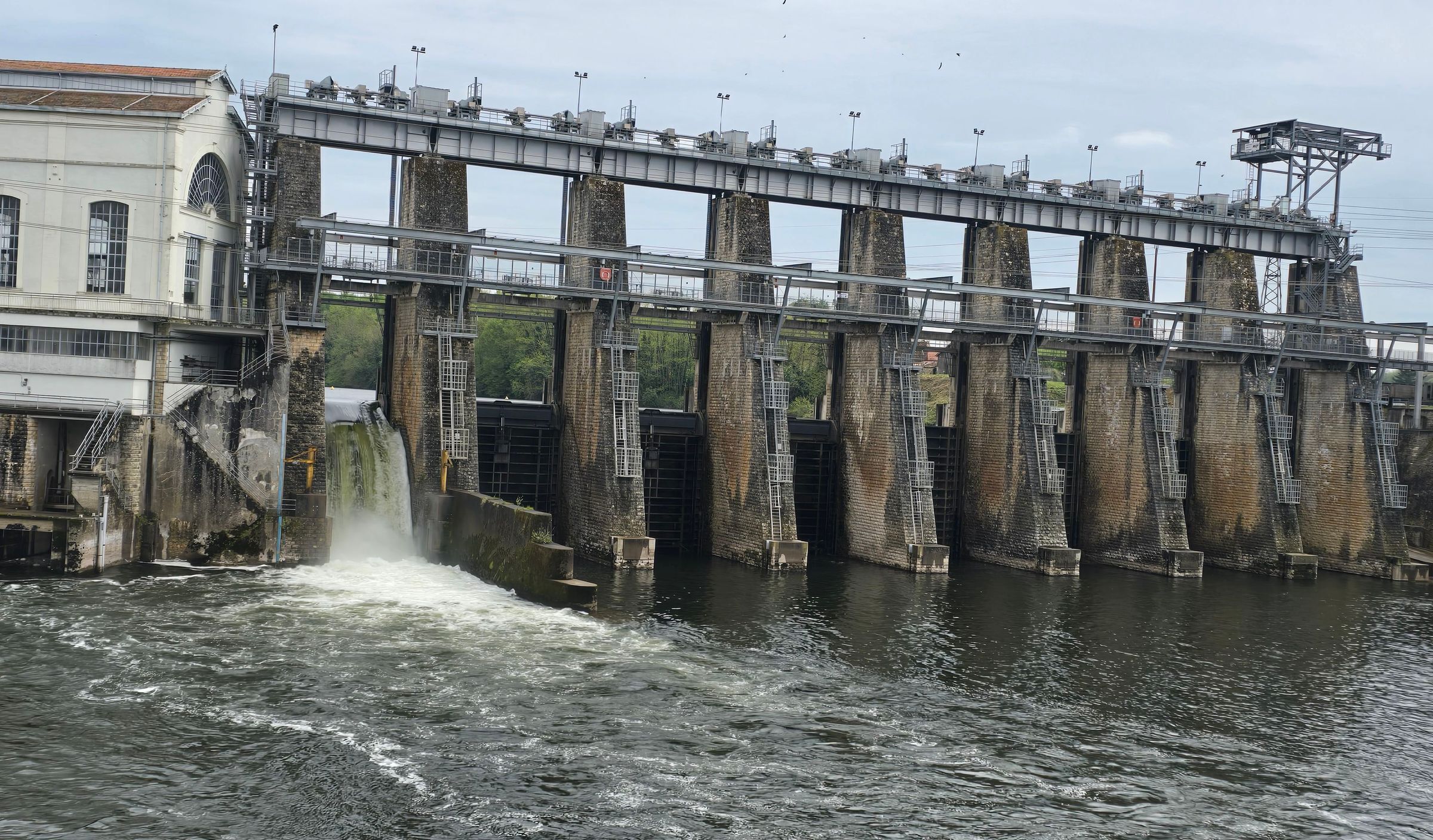 A dam with water flowing over it, a bridge, and a building with a staircase.