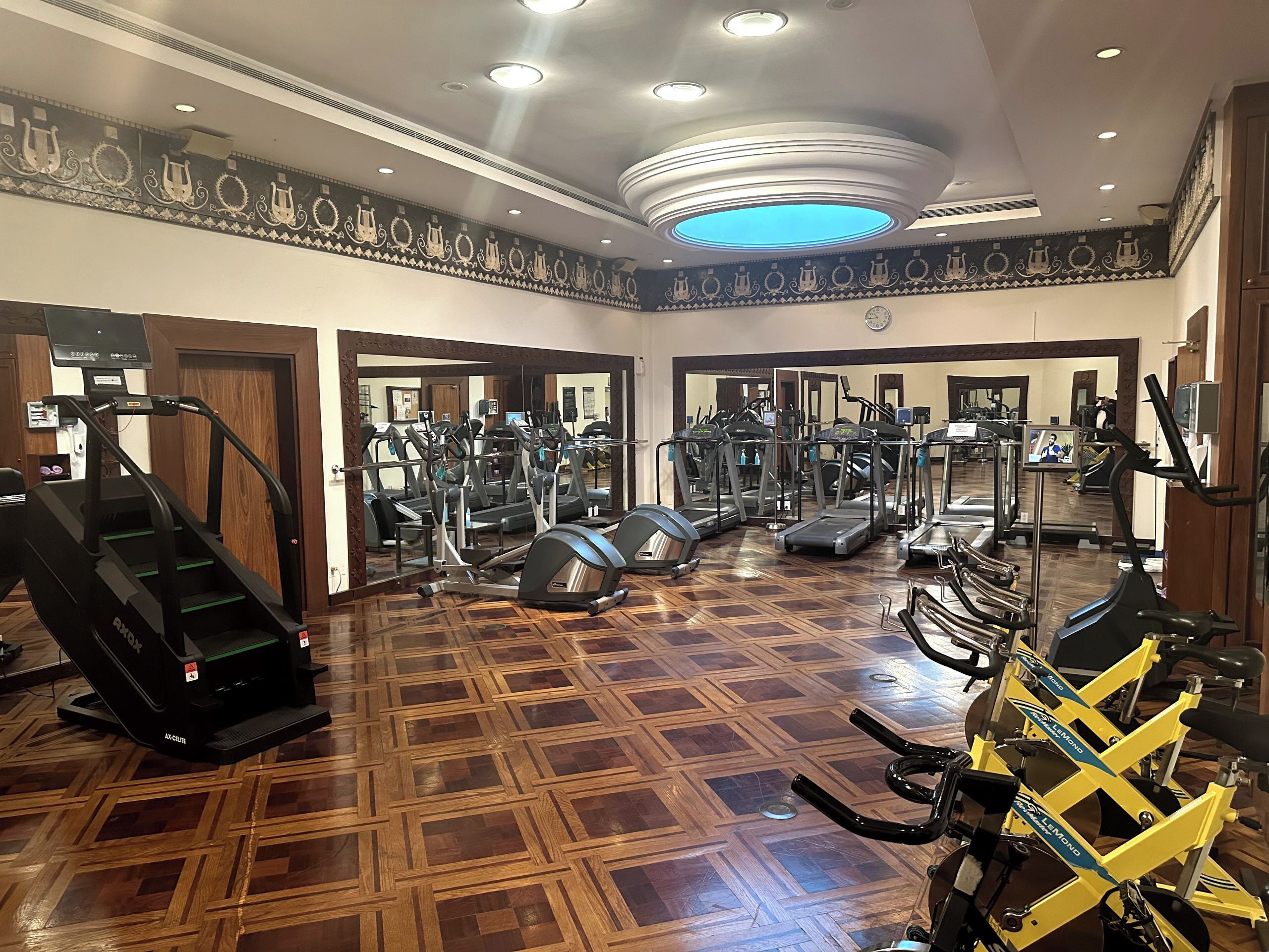 Une salle de sport équipée de tapis de course, de vélos elliptiques et d’appareils de musculation, dotée d’un sol en parquet et d’un mur recouvert de miroirs.