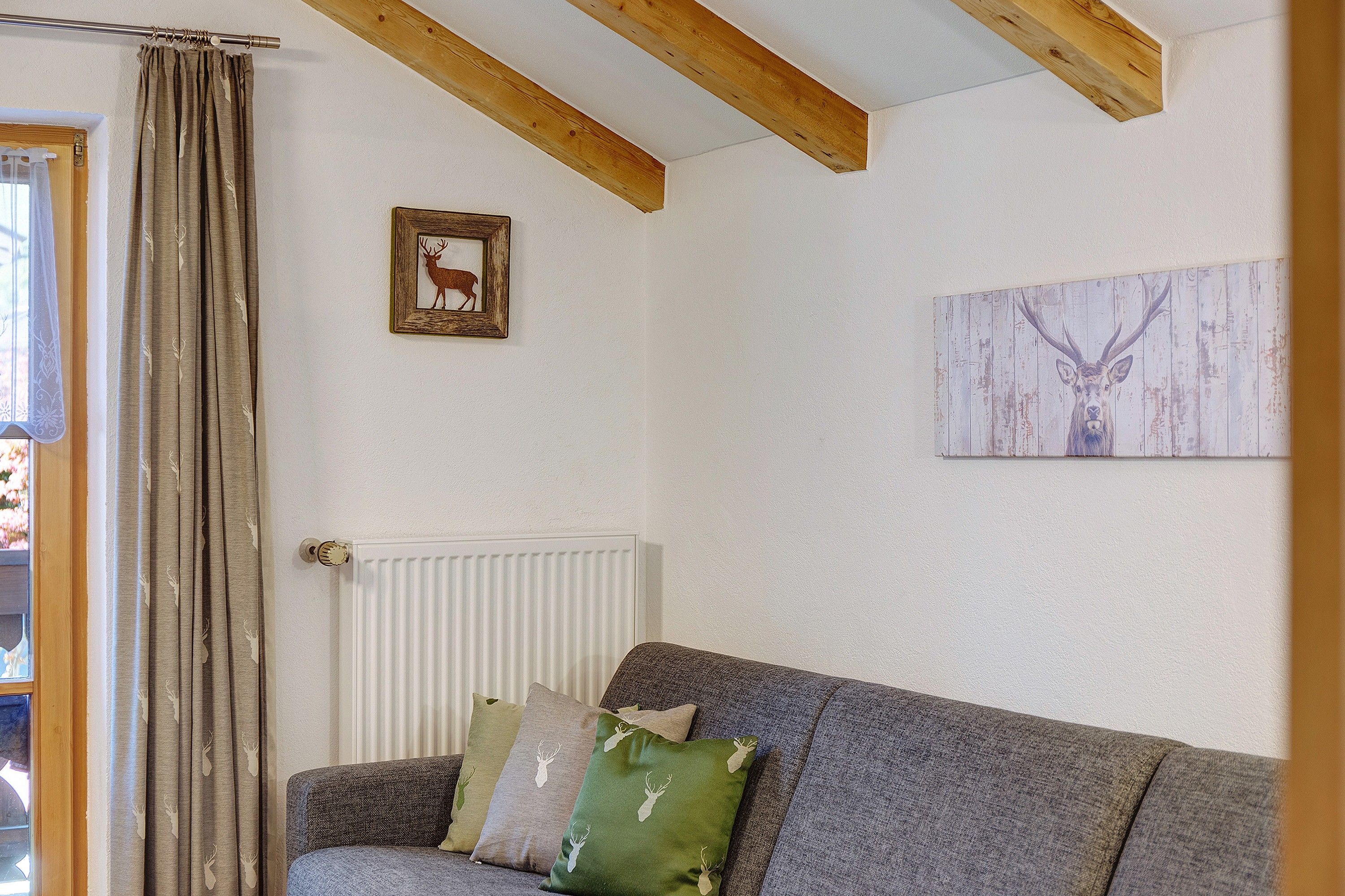 Een woonkamer met een grijze bank, een radiator, houten balken, een foto van een hert en een schilderij aan de muur.