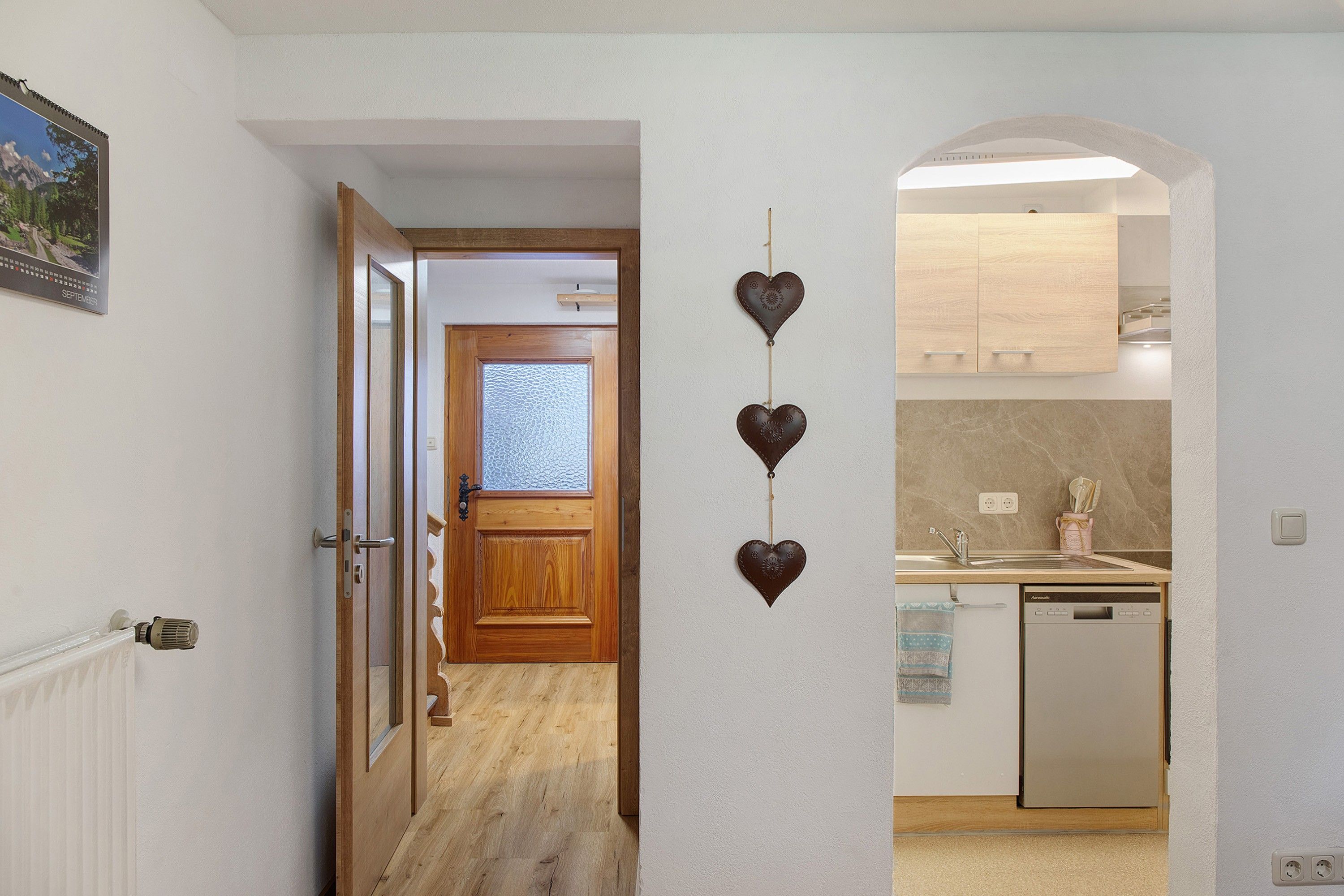 A hallway leading to a kitchen with a wooden door, a wall-mounted light fixture, and a calendar on the wall.