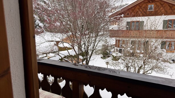 A wooden balcony with a view of a snow-covered house and trees.