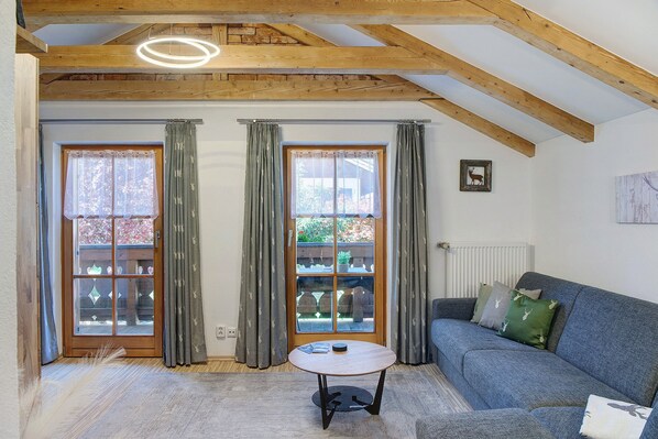 A room with a wooden ceiling, two windows with curtains, a sofa, a table, and a radiator.