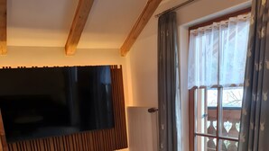 A room with a wooden floor, a large flat-screen TV mounted on the wall, a radiator, and a sliding glass door with curtains.