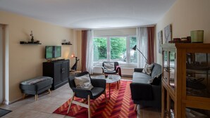 A living room with a sofa, armchair, and a small table. There is a television on a cabinet, a window with curtains, and a view of greenery outside. - Holiday Apartment "Zum Paladin" (Berg)