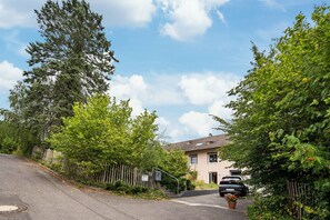 Une rue résidentielle avec une maison, une voiture garée devant, et un regard d’égout sur le trottoir.