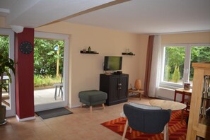 Uma sala de estar com televisão, um armário, uma poltrona, um tapete e uma porta que dá para uma varanda.