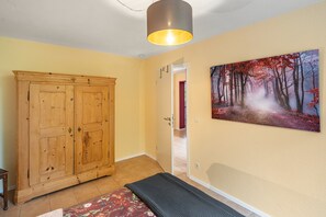Une pièce avec une armoire en bois, un banc, un tableau représentant une forêt et une porte donnant sur une autre pièce.